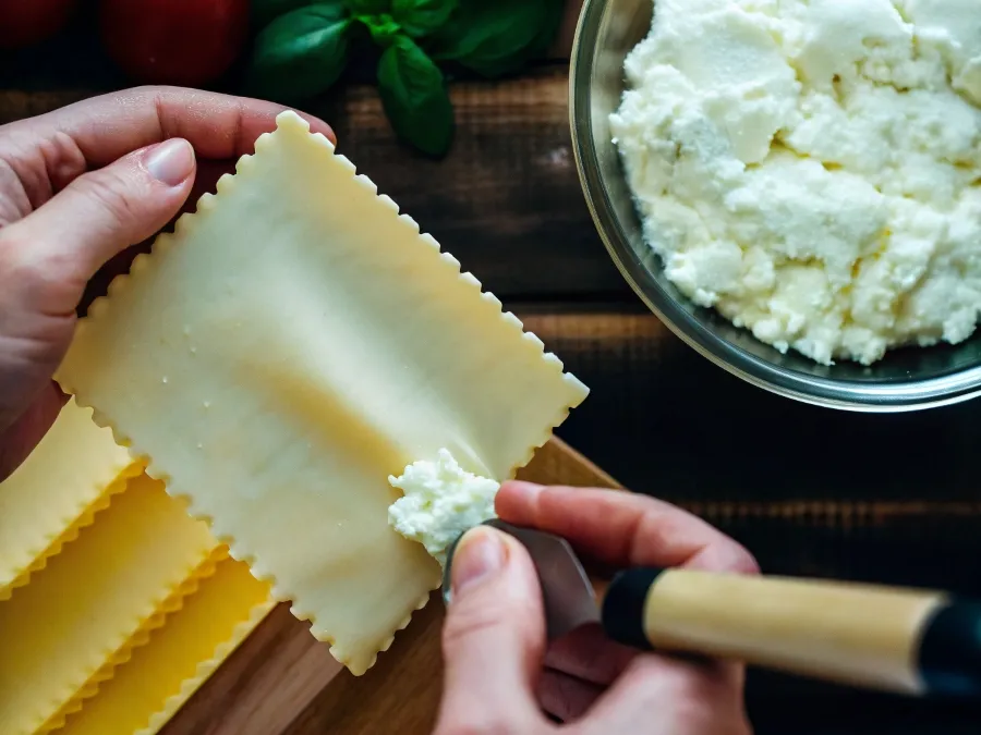 Spreading ricotta cheese onto lasagna sheets with fresh herbs and pasta.