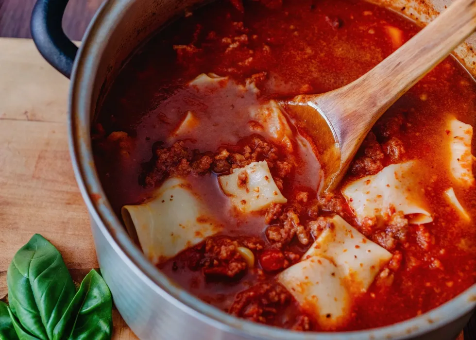 A simmering pot of lasagna soup with broken lasagna noodles, beef, and melted ricotta cheese, stirred with a wooden spoon.