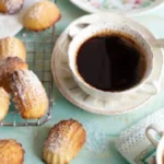 A cup of black coffee with freshly baked madeleines dusted with powdered sugar on a wire rack, set on a pastel blue table.