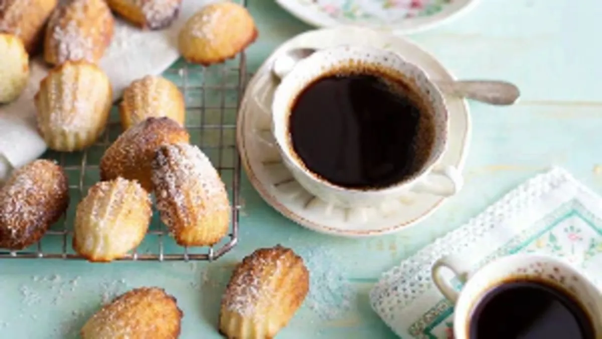 A cup of black coffee with freshly baked madeleines dusted with powdered sugar on a wire rack, set on a pastel blue table.