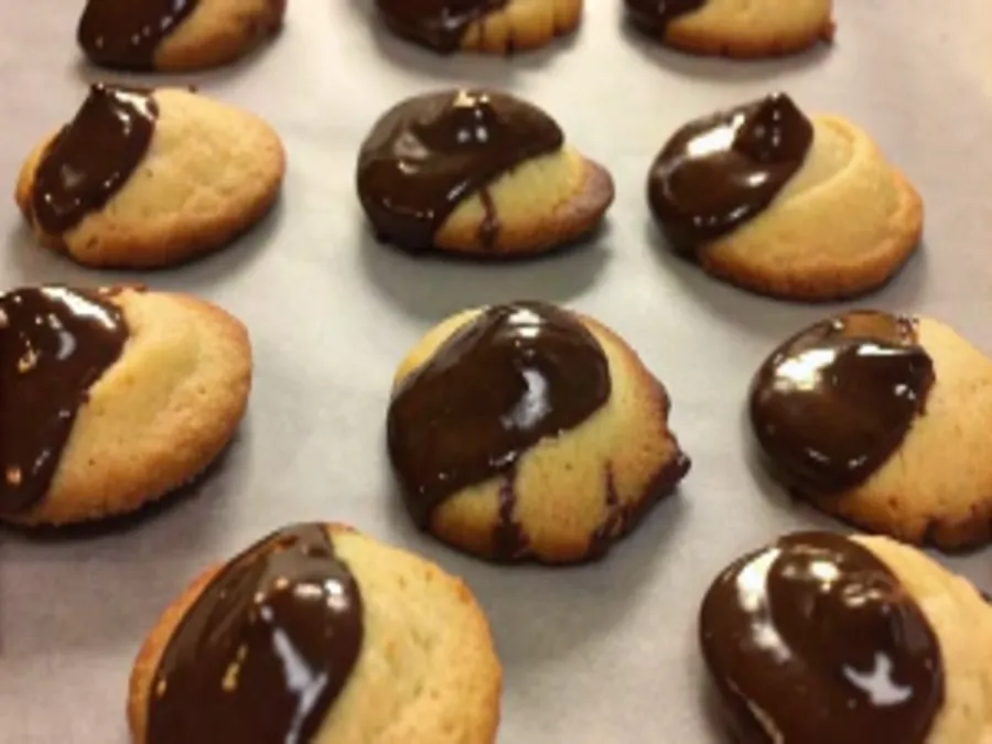 A batch of freshly baked madeleines partially dipped in glossy melted chocolate, arranged on a parchment-lined baking sheet.